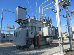 A large grey electrical transformer with metal pipes and cables in a substation