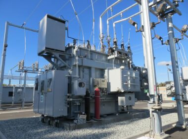 A large grey electrical transformer with metal pipes and cables in a substation