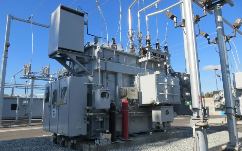 A large grey electrical transformer with metal pipes and cables in a substation