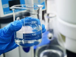 A gloved hand holding a glass beaker filled with clear liquid in a lab