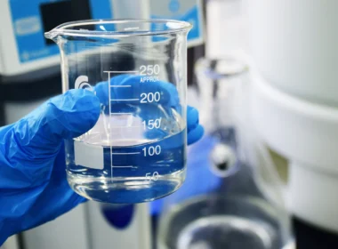 A gloved hand holding a glass beaker filled with clear liquid in a lab