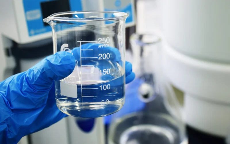 A gloved hand holding a glass beaker filled with clear liquid in a lab