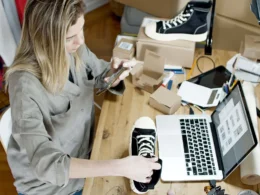 Small business owner photographing a sneaker for an online listing beside her laptop and packed orders.