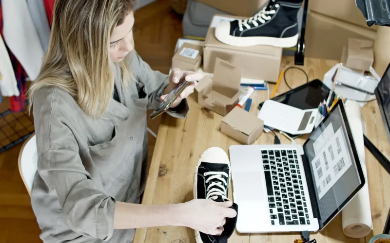 Small business owner photographing a sneaker for an online listing beside her laptop and packed orders.