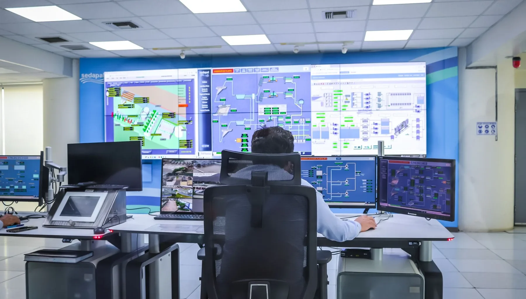 Operator at a desk in a control room facing a wall of system monitoring screens.