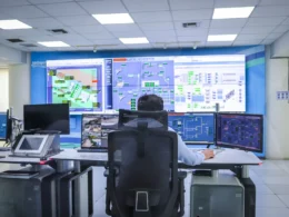 Operator at a desk in a control room facing a wall of system monitoring screens.