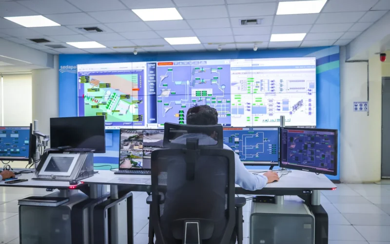 Operator at a desk in a control room facing a wall of system monitoring screens.