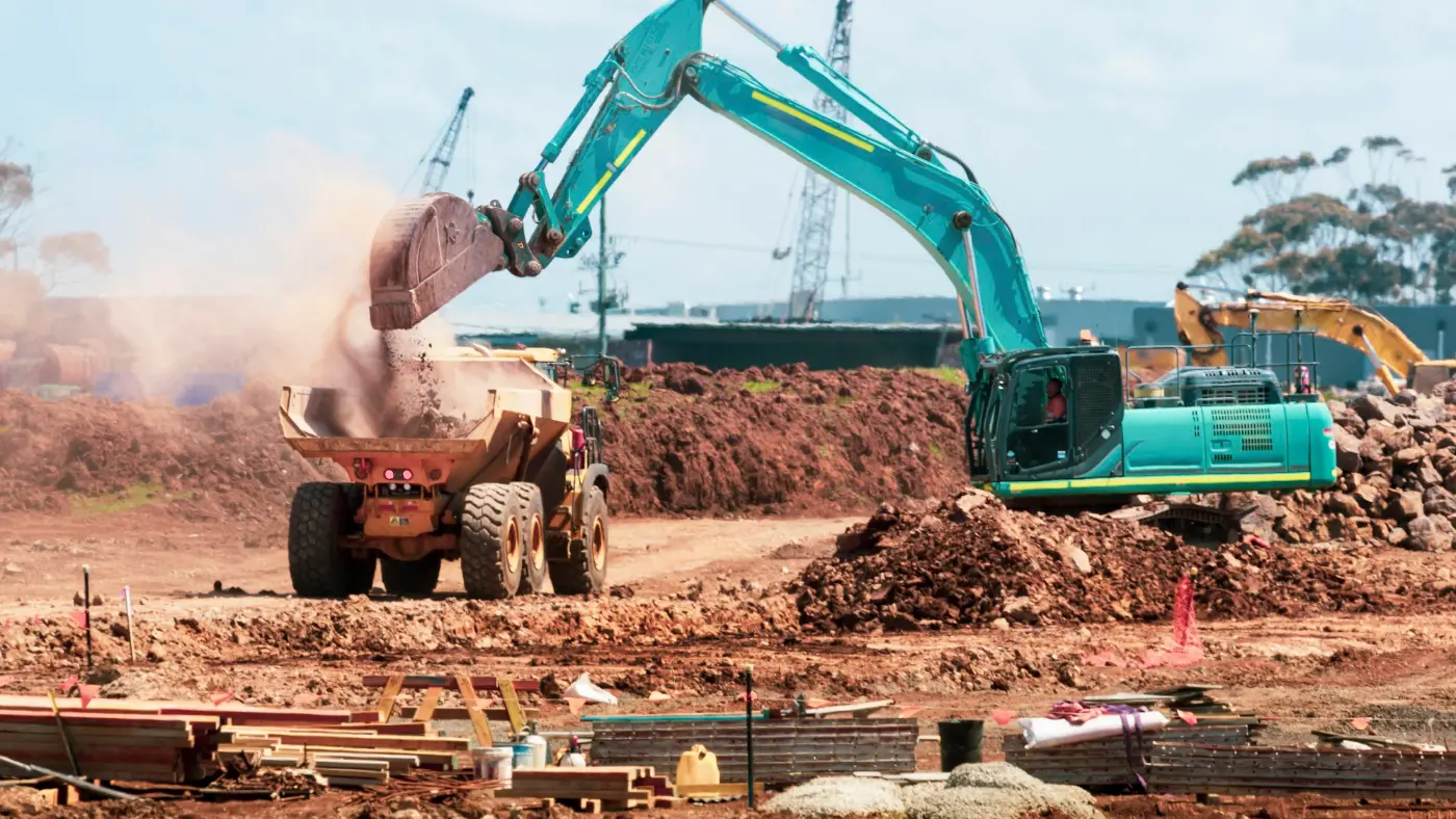 Teal excavator tipping soil into a yellow dump truck on a building site