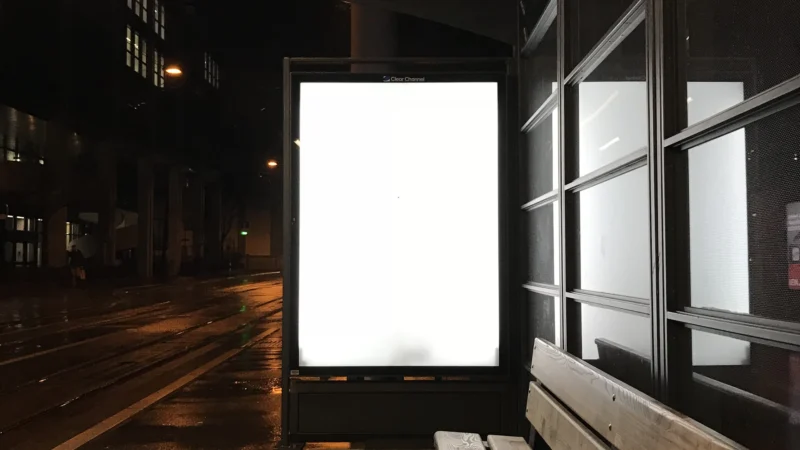 Blank glowing billboard at a wet bus stop at night