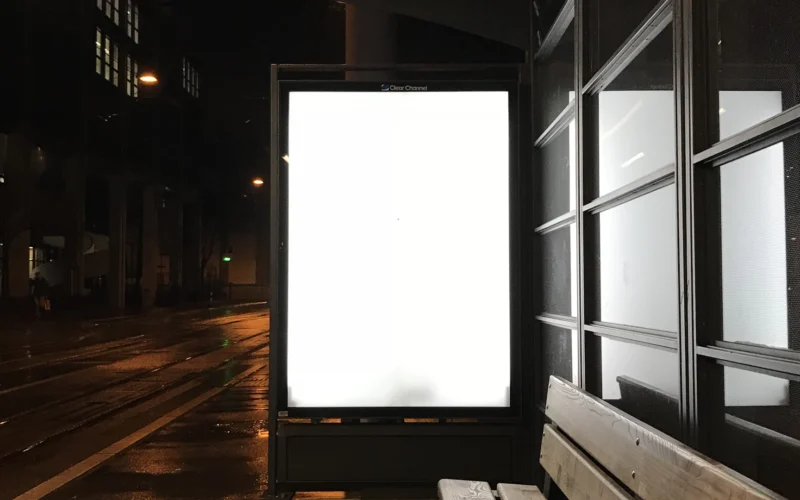 Blank glowing billboard at a wet bus stop at night