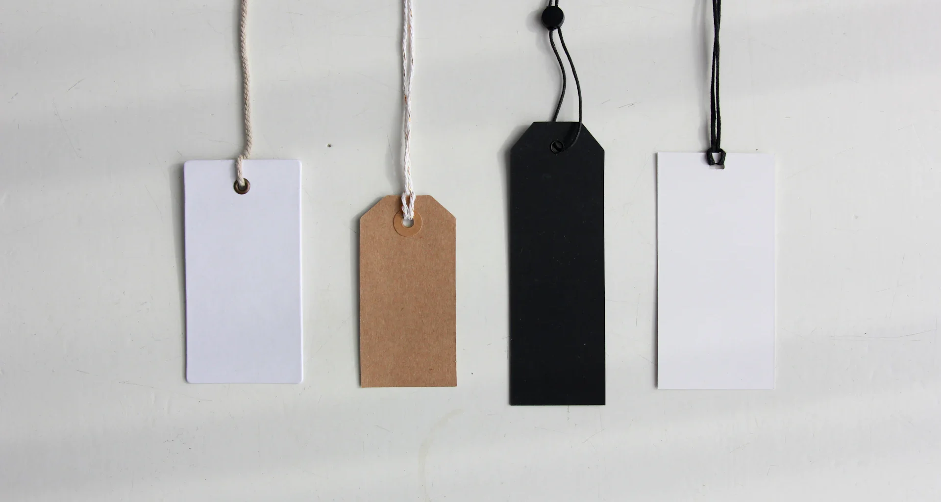 Four blank paper price tags in white, brown and black hanging on strings against a pale wall