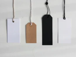 Four blank paper price tags in white, brown and black hanging on strings against a pale wall