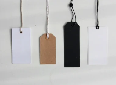 Four blank paper price tags in white, brown and black hanging on strings against a pale wall