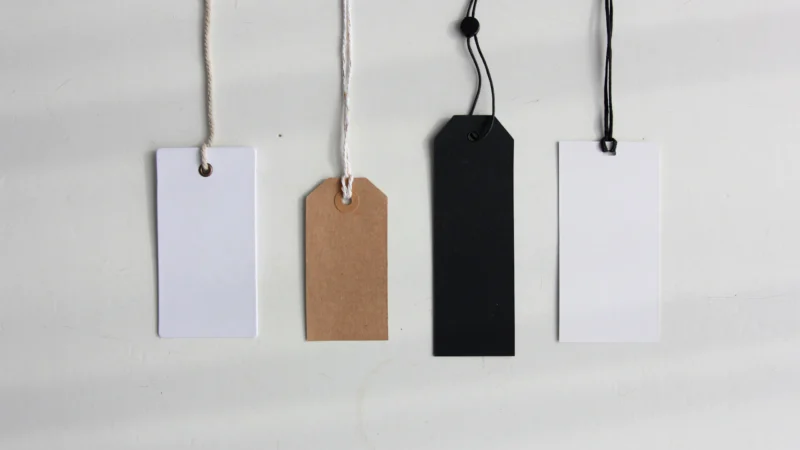 Four blank paper price tags in white, brown and black hanging on strings against a pale wall