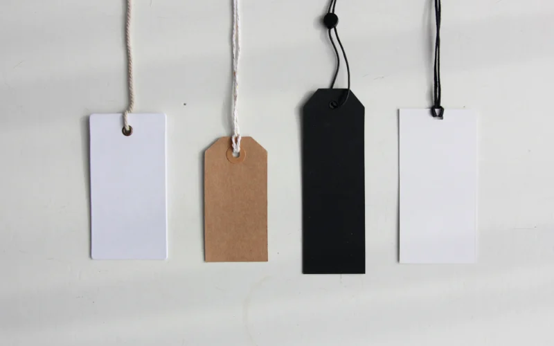 Four blank paper price tags in white, brown and black hanging on strings against a pale wall