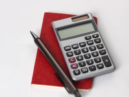 Silver calculator and black pen resting on a red notebook against a white background.