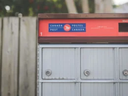 Red and grey Canada Post community mailbox beside a wooden fence, where an office's paper mail starts
