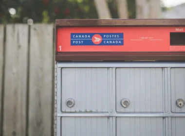 Red and grey Canada Post community mailbox beside a wooden fence, where an office's paper mail starts