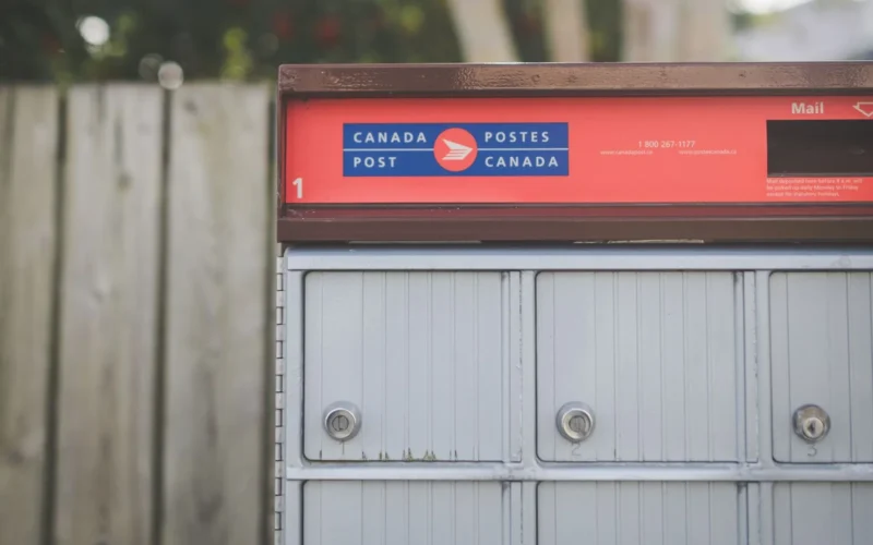 Red and grey Canada Post community mailbox beside a wooden fence, where an office's paper mail starts