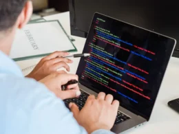 Developer typing colour-coded code on a laptop at an office desk.