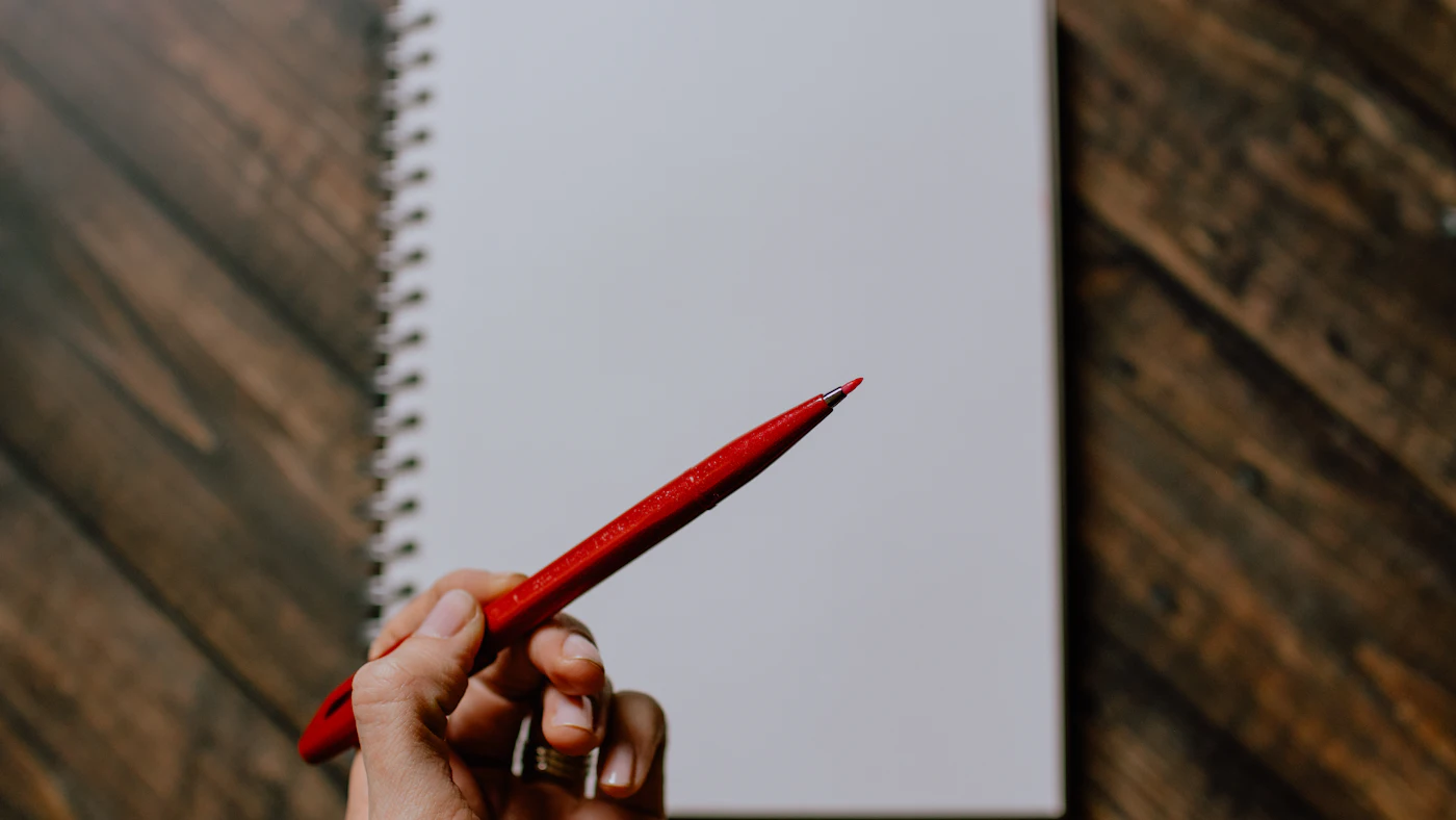 Hand holding a red pen over a blank spiral notebook on a wooden desk