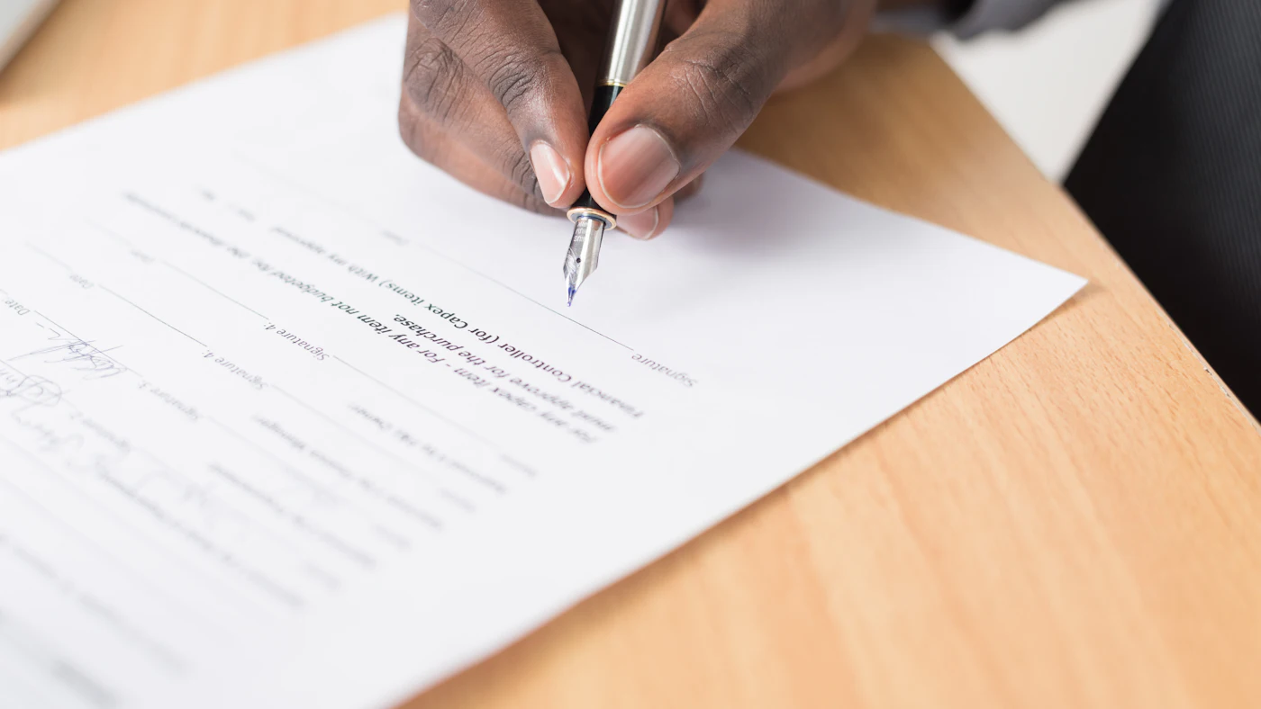 Hand signing a printed contract with a pen on a wooden desk