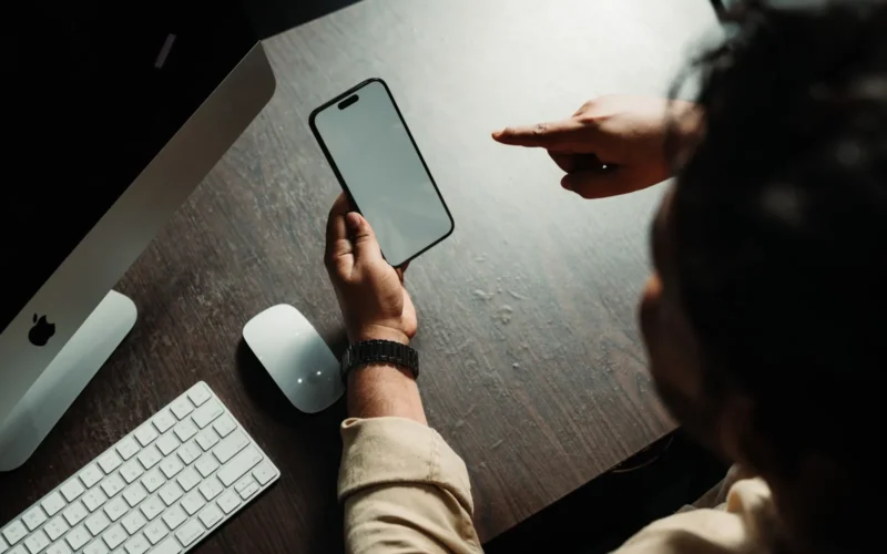 Person holding an iPhone at a desk beside a computer and keyboard, the setting where Siri AI meets work email