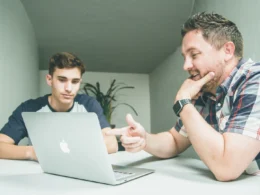 Two colleagues talking across a desk with a laptop in a bright office.