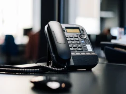 Black office desk phone with a coiled cord on a dark desk in an open-plan office