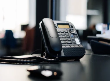 Black office desk phone with a coiled cord on a dark desk in an open-plan office