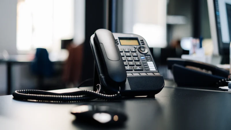 Black office desk phone with a coiled cord on a dark desk in an open-plan office
