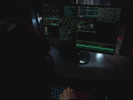 Person in a dark room at a desk with monitors full of scrolling code.