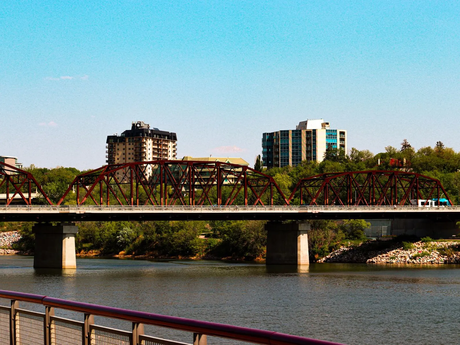Red steel bridge over the South Saskatchewan River with Saskatoon apartment towers behind it