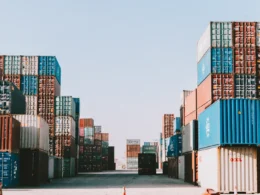 Stacks of shipping containers lining a port yard under a clear sky.