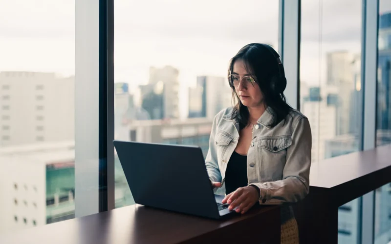Woman wearing headphones working on a laptop by a window